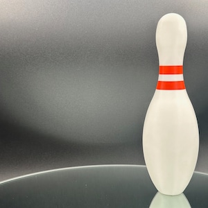 Puede incluir: Un pin de bolos blanco con dos rayas rojas alrededor del medio, de pie sobre una superficie de vidrio.