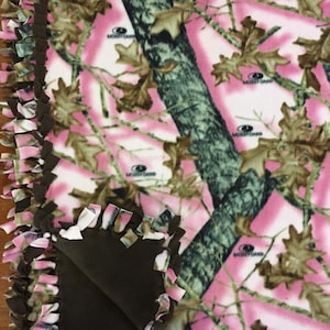 Op de afbeelding: Een roze en groene camouflage fleece deken met een bruine fleece achterkant. De deken heeft een geknoopte franje rand en de woorden "Mossy Oak" zijn op de stof gedrukt.