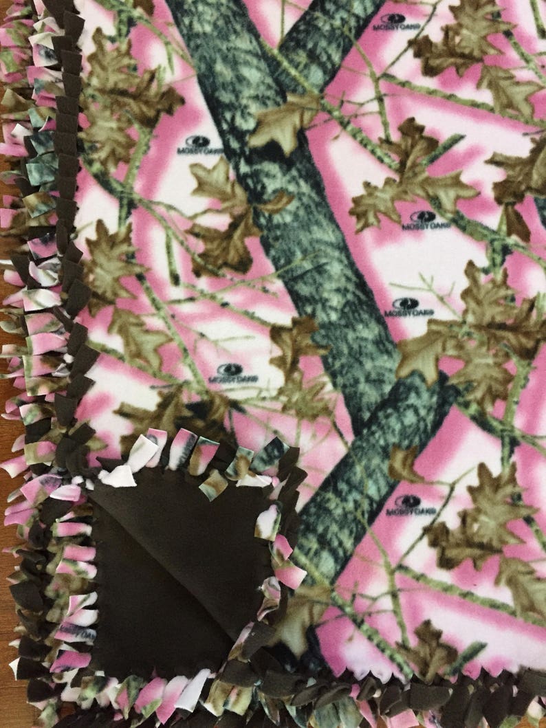 Op de afbeelding: Een roze en groene camouflage fleece deken met een bruine fleece achterkant. De deken heeft een geknoopte franje rand en de woorden "Mossy Oak" zijn op de stof gedrukt.