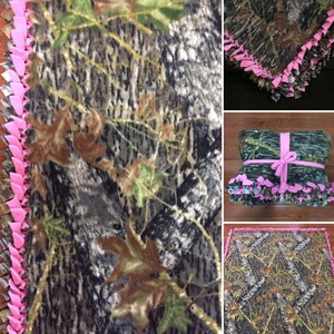 Op de afbeelding: Een camouflage fleece deken met een roze geknoopte franje rand. De deken is in drie&euml;n gevouwen en vastgebonden met een roze lint.