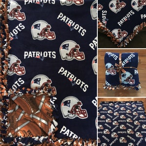 May include: A blue fleece blanket with a football helmet and the word "Patriots" repeated throughout. The blanket is tied with a brown ribbon and has a brown and white football pattern on the reverse side.
