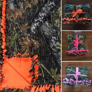 Op de afbeelding: Drie fleece-binddekens met een camouflagepatroon. De dekens zijn gebonden met een lint in verschillende kleuren: oranje, roze en roze.