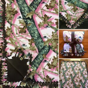 Op de afbeelding: Een roze en groene camouflage fleece deken met bruine banden. De deken is vastgebonden met een bruin lint en heeft de tekst "Mossy Oak" erop gedrukt.