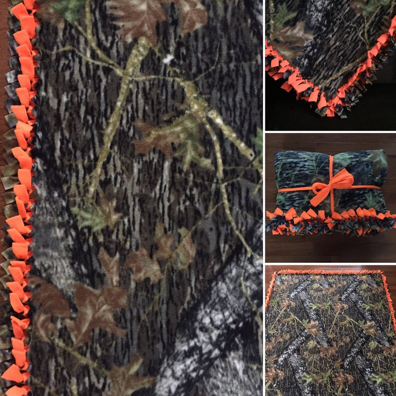 Op de afbeelding: Een camouflage fleece deken met een oranje geknoopte franje rand. De deken is gevouwen en in het midden vastgebonden met een lint.