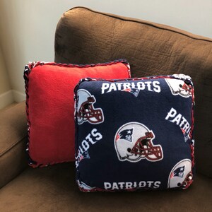 May include: Two decorative throw pillows, one red and one blue. The blue pillow has a repeated pattern of white football helmets and the word "Patriots" in red and white lettering.