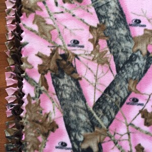 Op de afbeelding: Een roze en bruin camouflage fleece deken met een bruine geknoopte franje rand. De deken heeft de tekst "MOssy OAK" erop gedrukt.