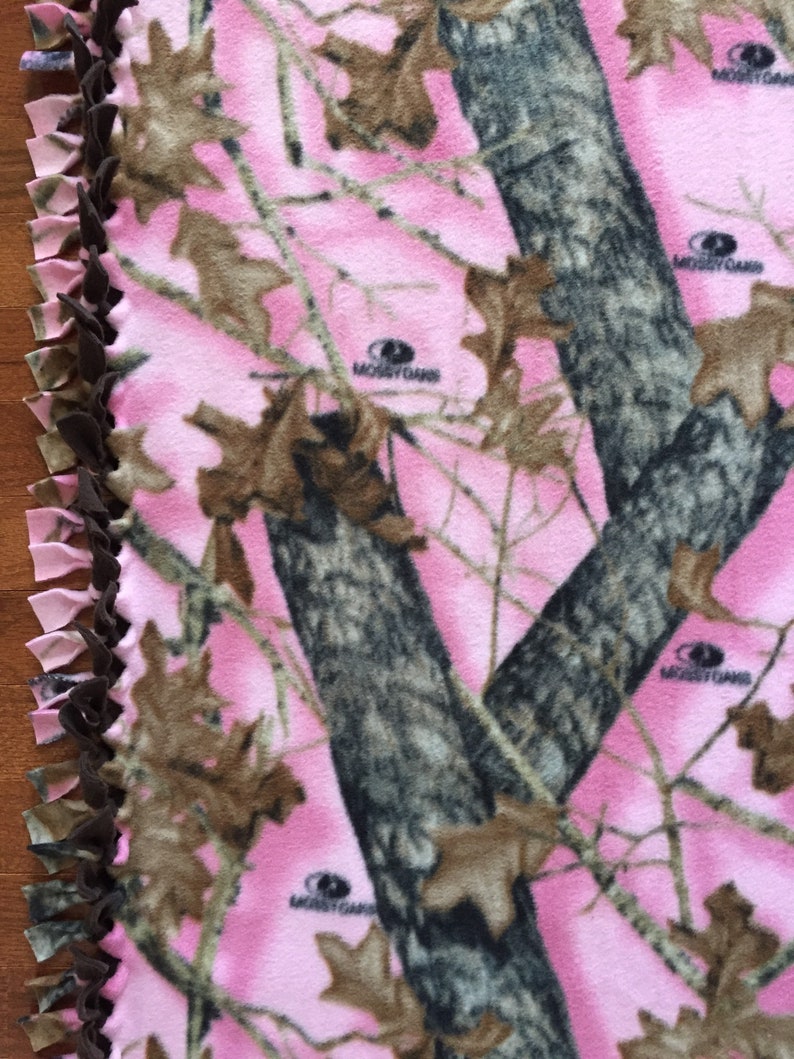 Op de afbeelding: Een roze en bruin camouflage fleece deken met een bruine geknoopte franje rand. De deken heeft de tekst "MOssy OAK" erop gedrukt.