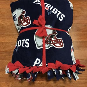 May include: A blue fleece blanket with a red ribbon tied around it. The blanket has a repeated pattern of football helmets and the word "Patriots" on it. The blanket has red fringe around the edges.