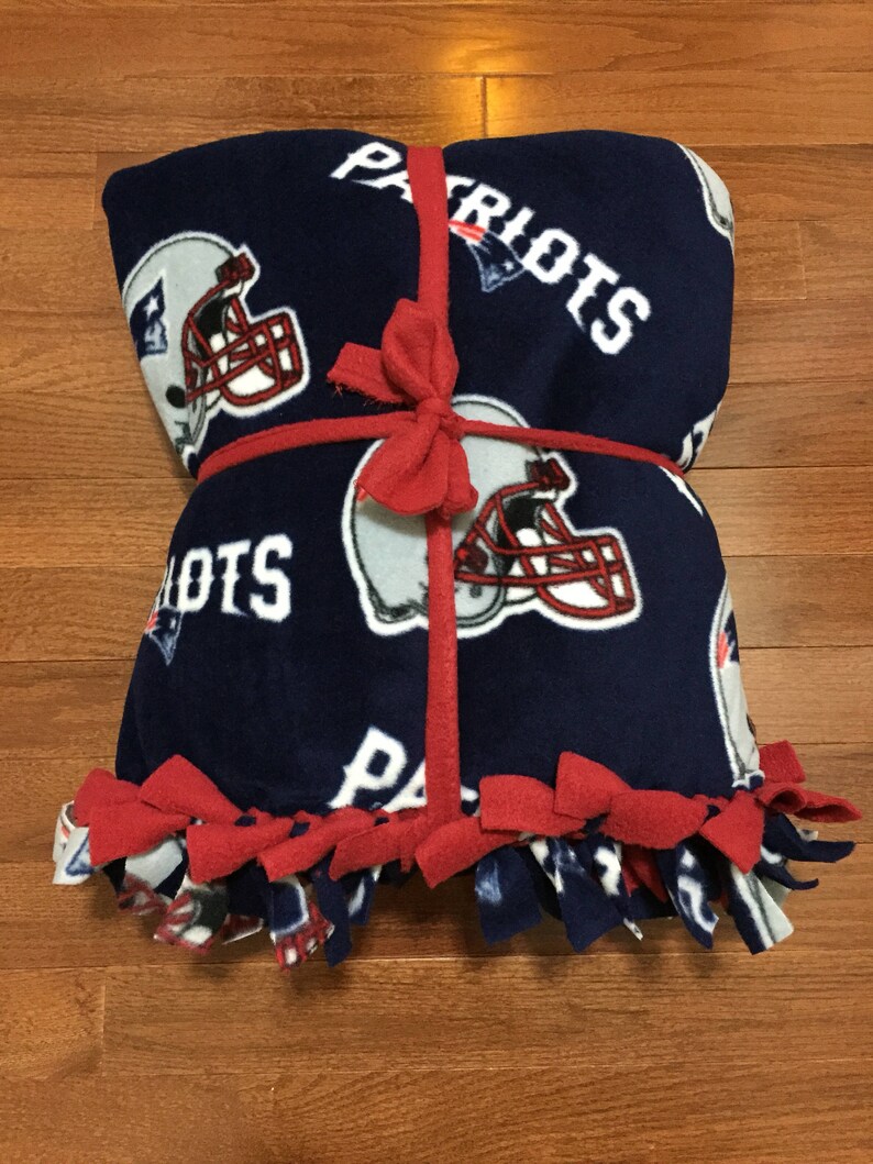 May include: A blue fleece blanket with a red ribbon tied around it. The blanket has a repeated pattern of football helmets and the word "Patriots" on it. The blanket has red fringe around the edges.