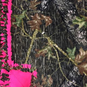 Op de afbeelding: Een roze en camouflage fleece-knoopdeken met een geknoopte franjerand. De deken is gemaakt van zacht, pluche stof en perfect om te knuffelen op de bank.