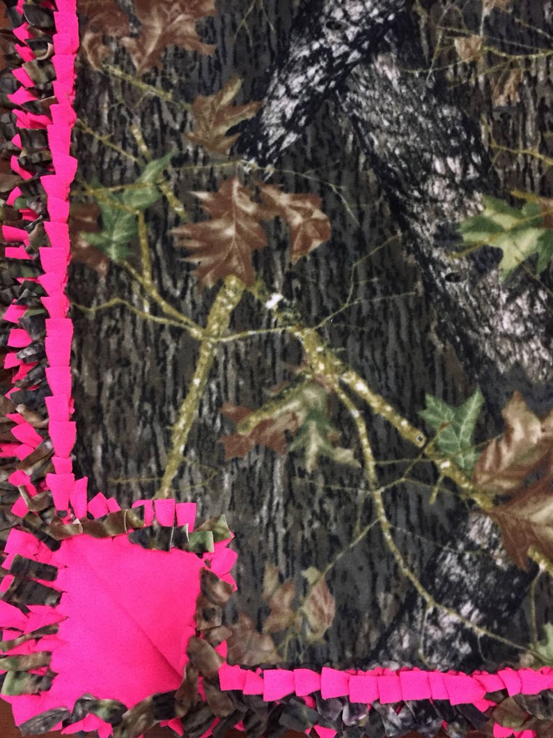 Op de afbeelding: Een roze en camouflage fleece-knoopdeken met een geknoopte franjerand. De deken is gemaakt van zacht, pluche stof en perfect om te knuffelen op de bank.