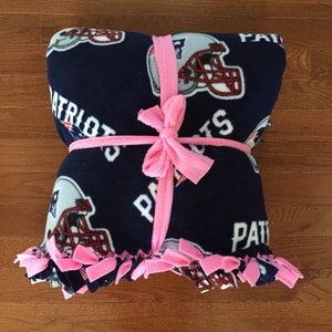 May include: A blue fleece blanket with a football helmet and the word "Patriots" printed on it. The blanket is tied with a pink ribbon.