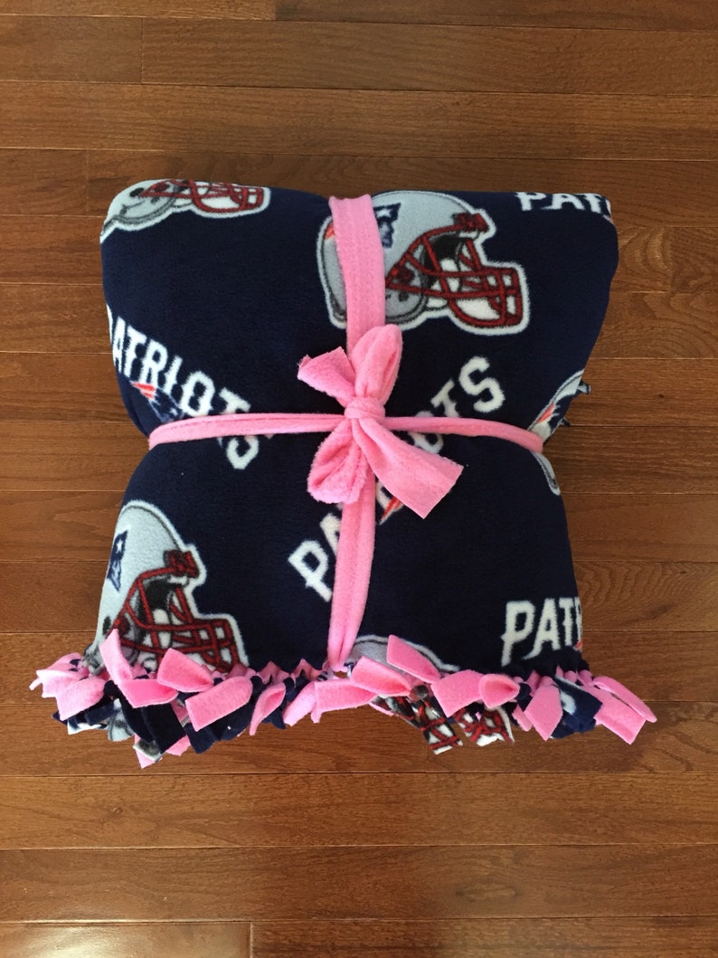 May include: A blue fleece blanket with a football helmet and the word "Patriots" printed on it. The blanket is tied with a pink ribbon.