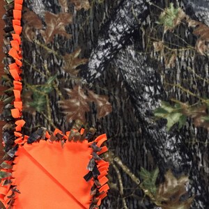 Op de afbeelding: Een camouflage fleece deken met een feloranje fleece rand en een groot oranje vierkant in de hoek. De deken is gemaakt met een geknoopte franje.