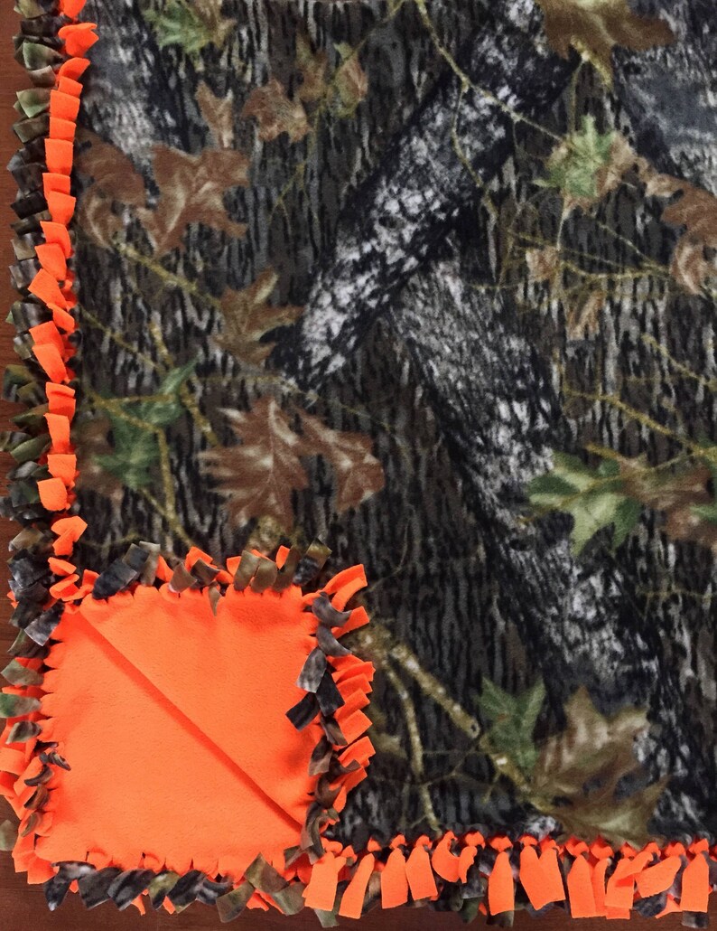 Op de afbeelding: Een camouflage fleece deken met een feloranje fleece rand en een groot oranje vierkant in de hoek. De deken is gemaakt met een geknoopte franje.
