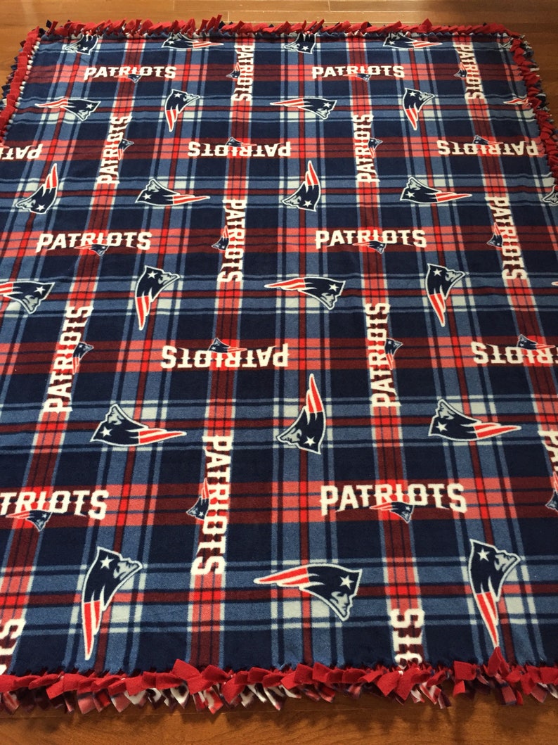 May include: A blue and red fleece blanket with a plaid pattern and the word "PATRIOTS" repeated throughout. The blanket has a knotted fringe border.
