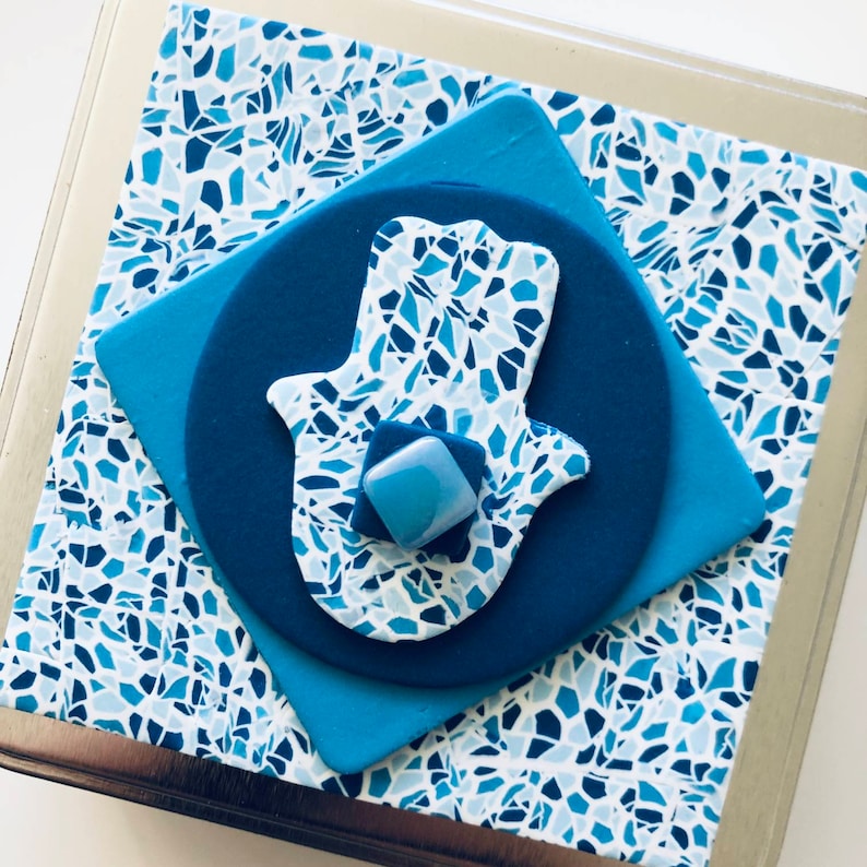 May include: A blue and white hamsa hand with a mosaic pattern, surrounded by a blue circle and square on a white background.