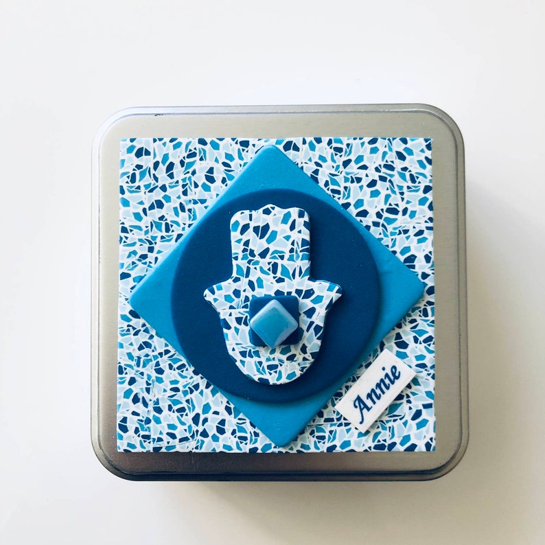 May include: A blue and white patterned metal tin with a hamsa hand charm and the name "Annie" on the front.