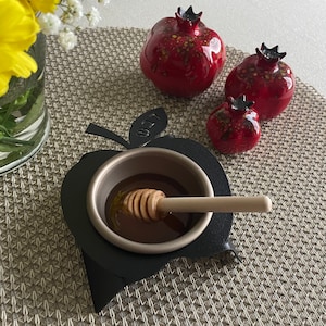 May include: A black metal honey dipper shaped like an apple with the Hebrew word for honey, "דבש" (dvash), inscribed on the side. The dipper is filled with honey and a wooden honey dipper is resting inside. Three red ceramic pomegranates are in the background.