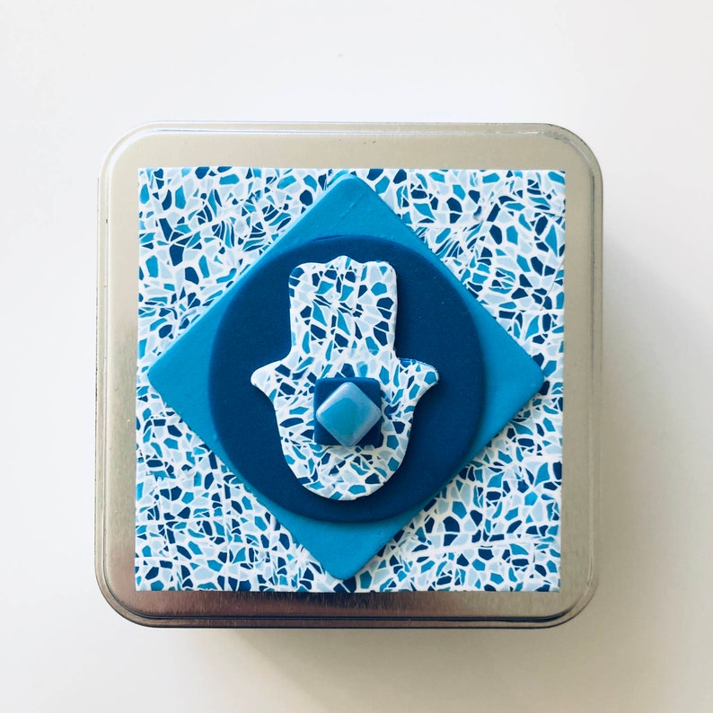 May include: A blue and white hamsa hand charm with a mosaic pattern, inside a square metal box.