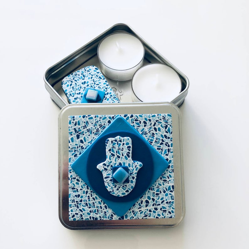 May include: A small metal tin with a blue and white mosaic design. The lid is open and reveals two white tea lights. The tin has a blue hamsa hand charm on the front.