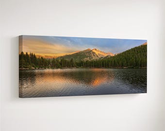 May include: A panoramic canvas print of a mountain lake at sunset. The image features a calm lake reflecting the sky's colors, with mountains and evergreen trees in the background. The sky transitions from blue to orange and yellow.