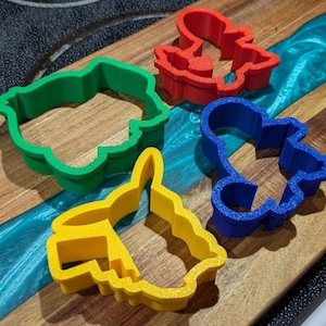 Puede incluir: Cuatro cortadores de galletas de colores rojo, verde, azul y amarillo, dispuestos sobre una tabla de cortar de madera. Los cortadores tienen forma de personajes de dibujos animados. La tabla tiene una veta de madera oscura con un río de resina azul.