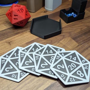 May include: A selection of gaming accessories, including a red D20 die, a black dice tray, a dice box, and several white and grey D20 coasters. The coasters have numbers from 0 to 20. The items are on a wooden surface.