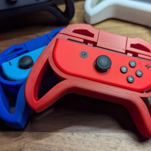 Op de afbeelding: Een close-up van verschillende game controller grips in verschillende kleuren. Een rode grip staat centraal, met een blauwe grip aan de linkerkant. De grips zijn ontworpen om game controllers vast te houden. De grips zijn gemaakt van plastic en hebben een getextureerd oppervlak.