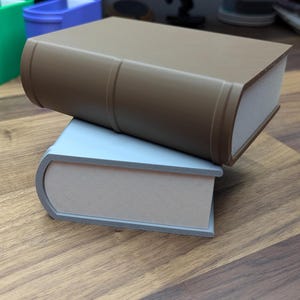 May include: Two decorative book-shaped boxes. The top box is brown, and the bottom box is light blue. Both boxes have a curved spine and a rectangular shape. The boxes are stacked on top of each other.