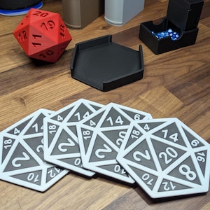 Puede incluir: Un conjunto de accesorios de juego, que incluye un dado D20 rojo, una bandeja para dados negra, una caja de dados y varios posavasos D20 blancos y grises. Los posavasos presentan n&uacute;meros del 0 al 20. Los art&iacute;culos est&aacute;n dispuestos sobre una superficie de madera.