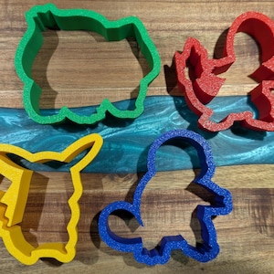 Puede incluir: Cuatro cortadores de galletas de colores con forma de personajes de dibujos animados. Los cortadores son verdes, rojos, amarillos y azules. Los cortadores est&aacute;n sobre una superficie de madera con un r&iacute;o de epoxi azul y verde.