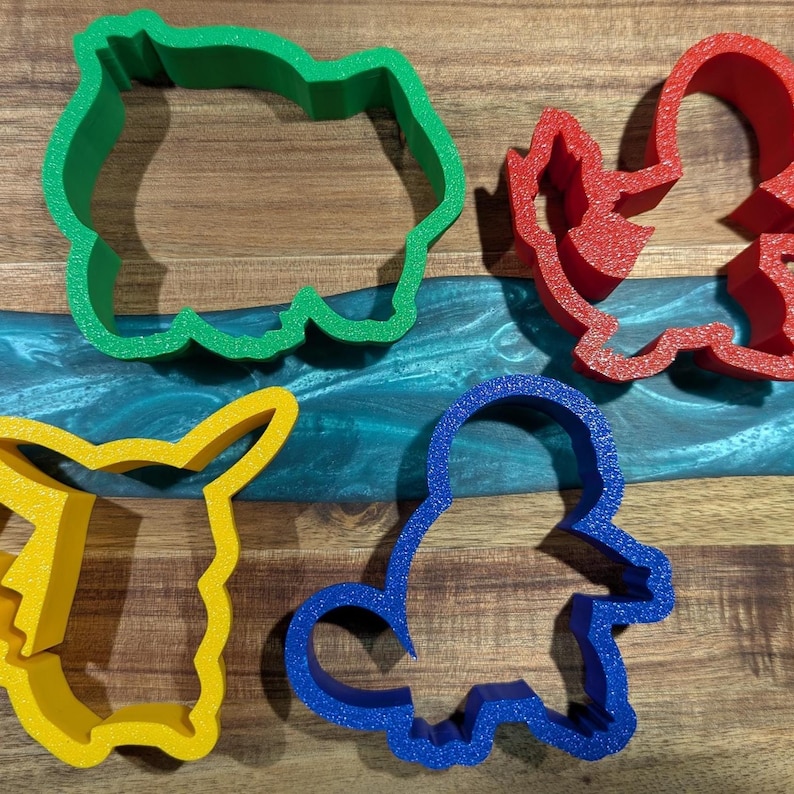 Puede incluir: Cuatro cortadores de galletas de colores con forma de personajes de dibujos animados. Los cortadores son verdes, rojos, amarillos y azules. Los cortadores est&aacute;n sobre una superficie de madera con un r&iacute;o de epoxi azul y verde.