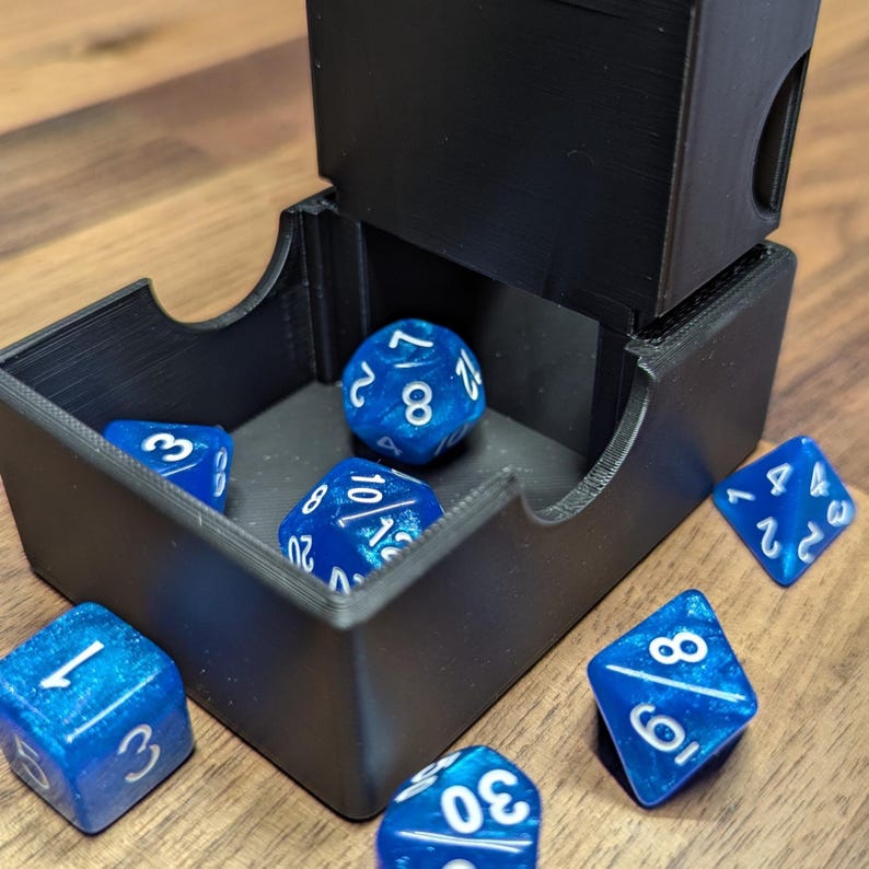Puede incluir: Una torre de dados negra con una bandeja, que contiene varios dados azules y blancos. Los dados est&aacute;n esparcidos alrededor de la torre sobre una superficie de madera. Los dados tienen n&uacute;meros blancos.