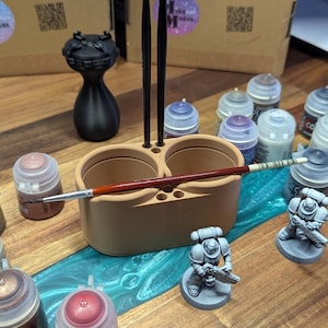 Puede incluir: Un soporte beige para pinceles con dos compartimentos y varios pinceles. Figuras en miniatura y botes de pintura de varios colores rodean el soporte. La escena está ambientada sobre una superficie de madera con un río de epoxi azul y verde.