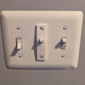 Puede incluir: Una placa de interruptor de luz blanca rectangular con tres interruptores. Dos est&aacute;n apagados, uno est&aacute; marcado "ON". La placa est&aacute; montada en una pared gris.