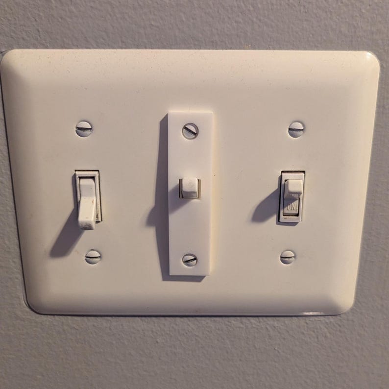 Puede incluir: Una placa de interruptor de luz blanca rectangular con tres interruptores. Dos est&aacute;n apagados, uno est&aacute; marcado "ON". La placa est&aacute; montada en una pared gris.