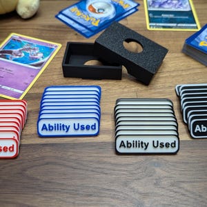 May include: A collection of trading card game accessories, including card stacks, a black card holder, and several "Ability Used" markers in red, blue, black, and white. The cards feature various designs and logos.