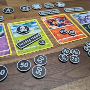 Puede incluir: Un conjunto de cartas coleccionables de Pok&eacute;mon con fichas y marcadores de juego. Las cartas presentan ilustraciones coloridas y texto, con fichas circulares que muestran n&uacute;meros como 20 y 50. Los marcadores rectangulares dicen "Ability Used".