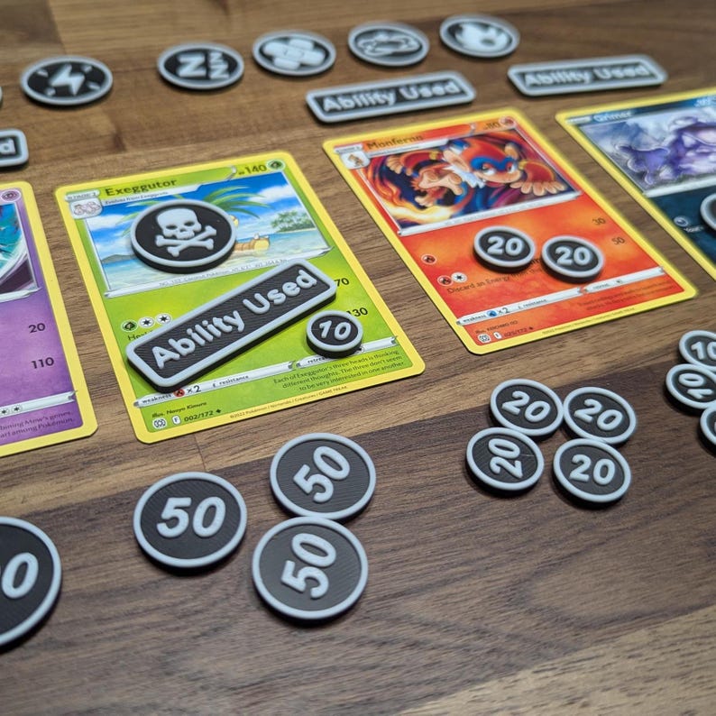 Puede incluir: Un conjunto de cartas coleccionables de Pok&eacute;mon con fichas y marcadores de juego. Las cartas presentan ilustraciones coloridas y texto, con fichas circulares que muestran n&uacute;meros como 20 y 50. Los marcadores rectangulares dicen "Ability Used".