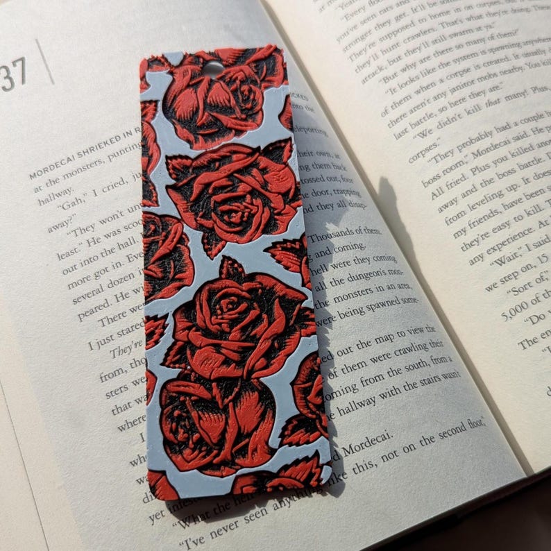 Puede incluir: Marcap&aacute;ginas rectangular con un patr&oacute;n repetido de rosas rojas sobre un fondo azul claro. El marcap&aacute;ginas est&aacute; hecho de un material resistente y tiene un agujero en la parte superior para una borla. El dise&ntilde;o es detallado y llamativo.
