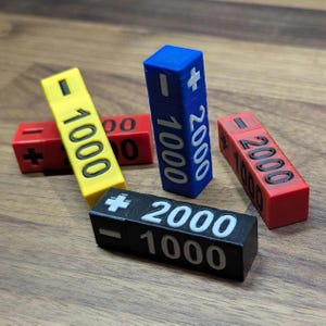 Puede incluir: Un conjunto de bloques rectangulares coloridos con números y símbolos impresos. Los bloques son amarillos, rojos, azules y negros, con texto blanco que indica valores como "1000" y "2000", junto con símbolos de más y menos. Los bloques están dispuestos sobre una superficie de madera.
