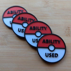 Puede incluir: Cuatro fichas circulares rojas, blancas y negras con las palabras "ABILITY" y "USED" impresas. Las fichas est&aacute;n dispuestas en diagonal sobre una superficie de madera marr&oacute;n claro. El dise&ntilde;o se asemeja a una popular serie de animaci&oacute;n.