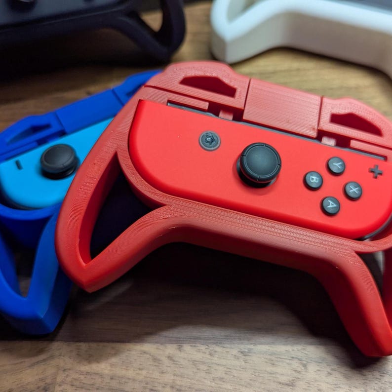 Puede incluir: Primer plano de varios mandos de juego en varios colores, incluyendo rojo, azul y negro. Los mandos tienen una superficie texturizada y cuentan con botones y joysticks. El mando rojo est&aacute; enfocado, con los otros mandos ligeramente borrosos en el fondo.