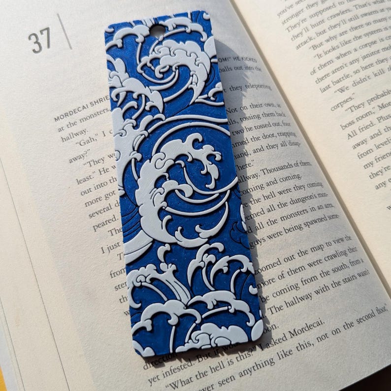 Puede incluir: Marcap&aacute;ginas azul y blanco con dise&ntilde;o de olas. El marcap&aacute;ginas tiene un agujero en la parte superior para una borla. El dise&ntilde;o es un patr&oacute;n repetitivo de olas estilizadas. El marcap&aacute;ginas est&aacute; sobre las p&aacute;ginas de un libro.