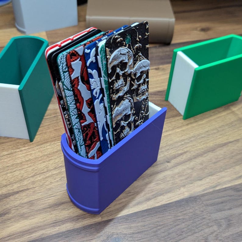 Puede incluir: Un soporte morado con forma de libro que contiene varios marcadores decorativos. Los marcadores presentan varios dise&ntilde;os, incluyendo rosas, patrones abstractos y calaveras. El soporte y los marcadores est&aacute;n sobre una superficie de madera.