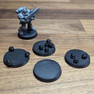 Puede incluir: Una figura en miniatura gris de un Space Marine con un arma, sobre una base negra. Cuatro bases circulares negras cercanas, algunas con peque&ntilde;as decoraciones de calaveras, para juegos de mesa.