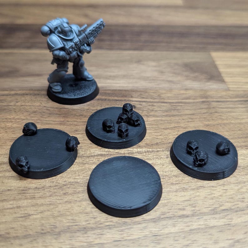Puede incluir: Una figura en miniatura gris de un Space Marine con un arma, sobre una base negra. Cuatro bases circulares negras cercanas, algunas con peque&ntilde;as decoraciones de calaveras, para juegos de mesa.