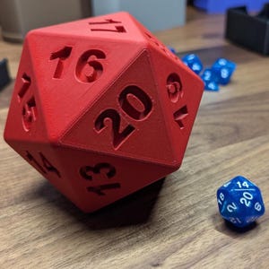 May include: A large red twenty-sided die with black numbers, next to a smaller blue twenty-sided die. The large die is the focal point, with the number 20 prominently displayed. Several other blue dice are visible in the background. The dice are likely used for tabletop gaming.