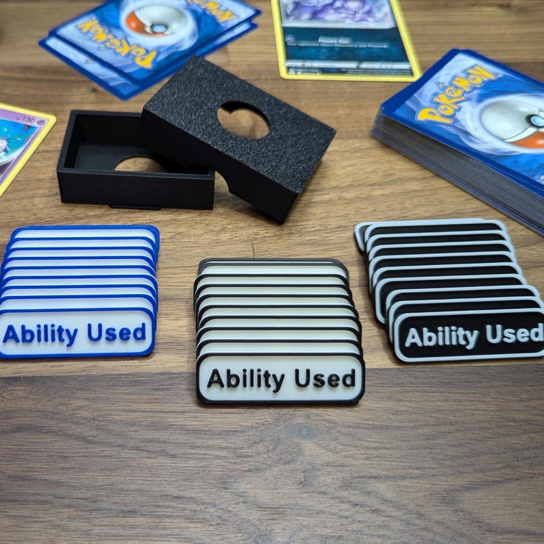 Puede incluir: Un tarjetero negro y tres fichas "Ability Used" en azul, negro y blanco y negro. Varias cartas coleccionables son visibles en el fondo. Las fichas tienen un dise&ntilde;o a rayas y el texto "Ability Used".